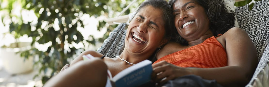 A mother and daughter lie together in a hammock surrounded by lush greenery, with the mother wearing an orange top and holding a book, while the daughter laughs while resting her head on her mother's shoulder.