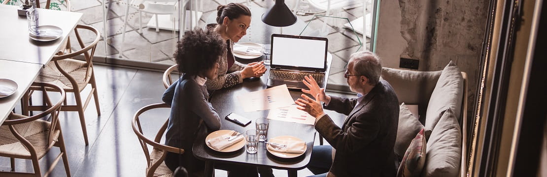 An overhead shot of three professionals sitting at a café table together. A few papers and an open laptop are visible on the table.